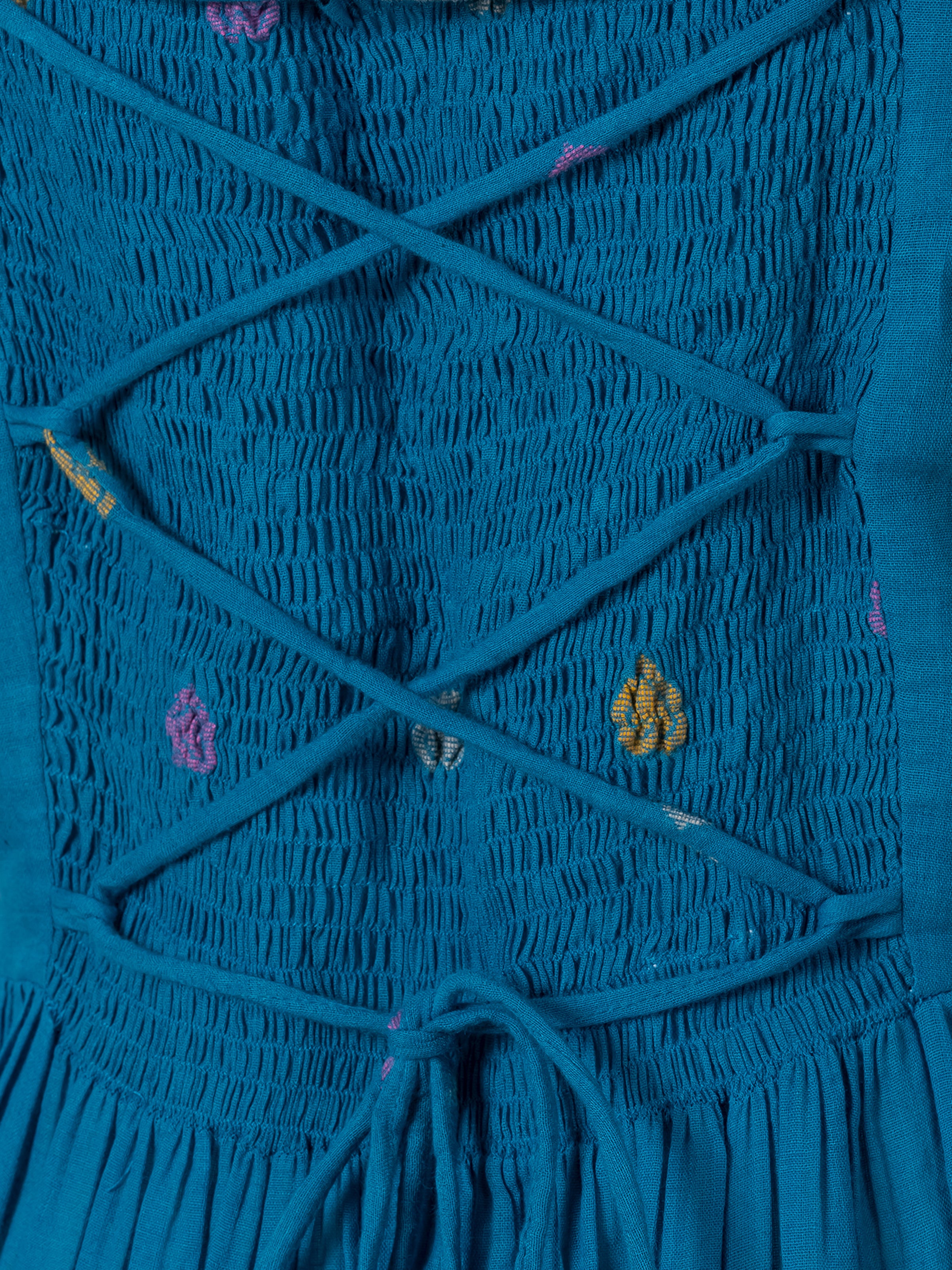 Teal cotton maxi dress with smocked texture and colorful polka dot embroidery, close-up back view showing crisscross straps and tie detail