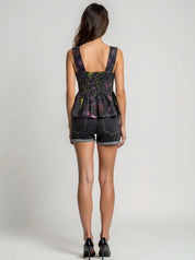 Woman wearing a black sleeveless top with colorful patterns and denim shorts on a gray background