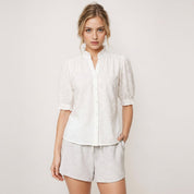 Woman wearing a white blouse with floral patterns and light gray shorts on a plain background